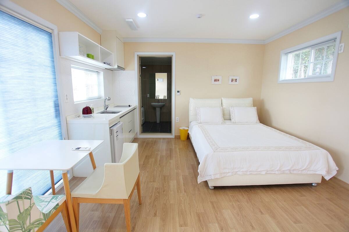 A bedroom with a bed, a desk, and a chair. There is a window with blinds and a door leading to a bathroom.