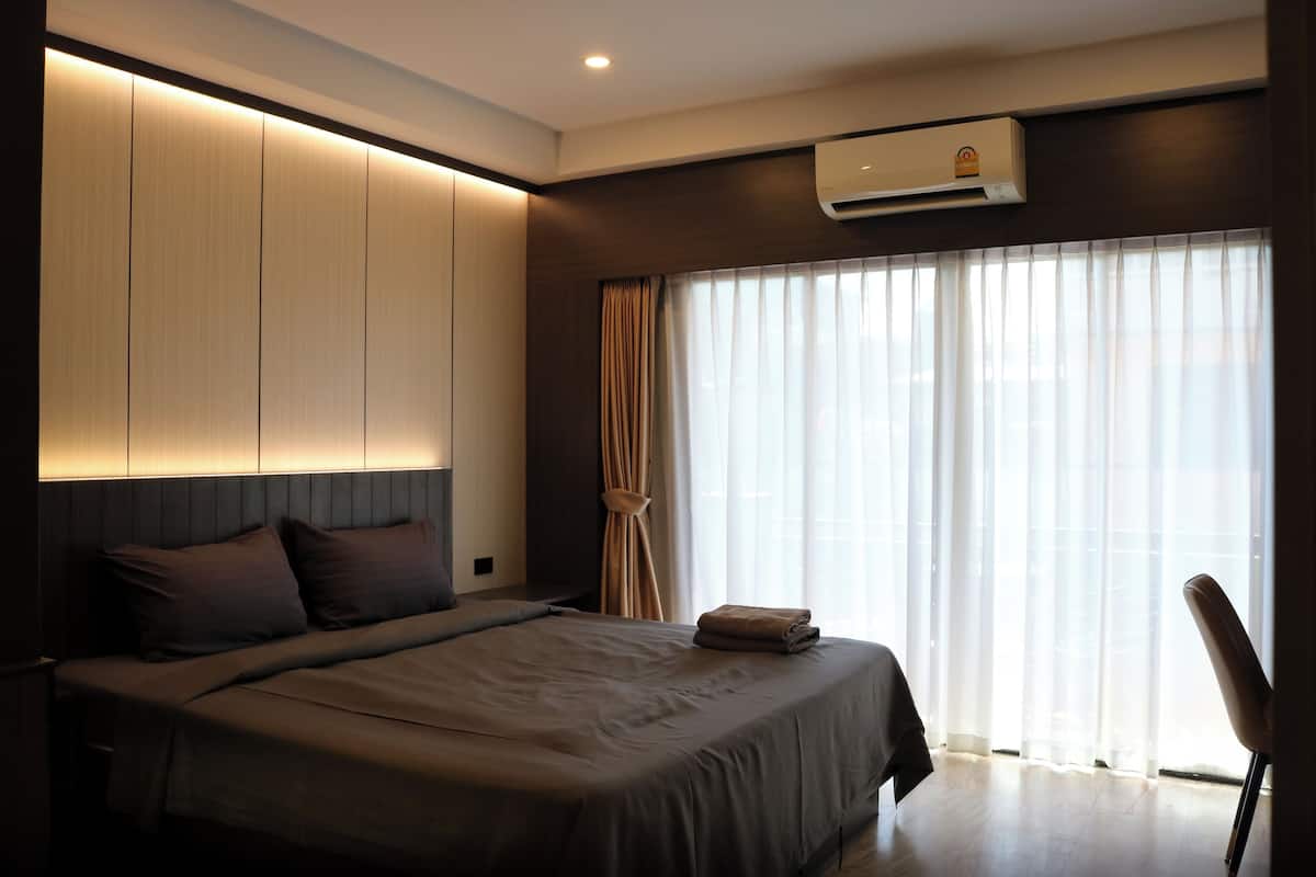 A modern hotel room with a large bed, a built-in wardrobe, an air conditioning unit, and a large window with sheer curtains.