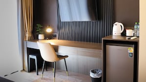 A modern hotel room with a wooden desk, a chair, a flat-screen TV mounted on the wall, a small kitchenette with a kettle, and a trash bin. - Max Residence (Pattaya)