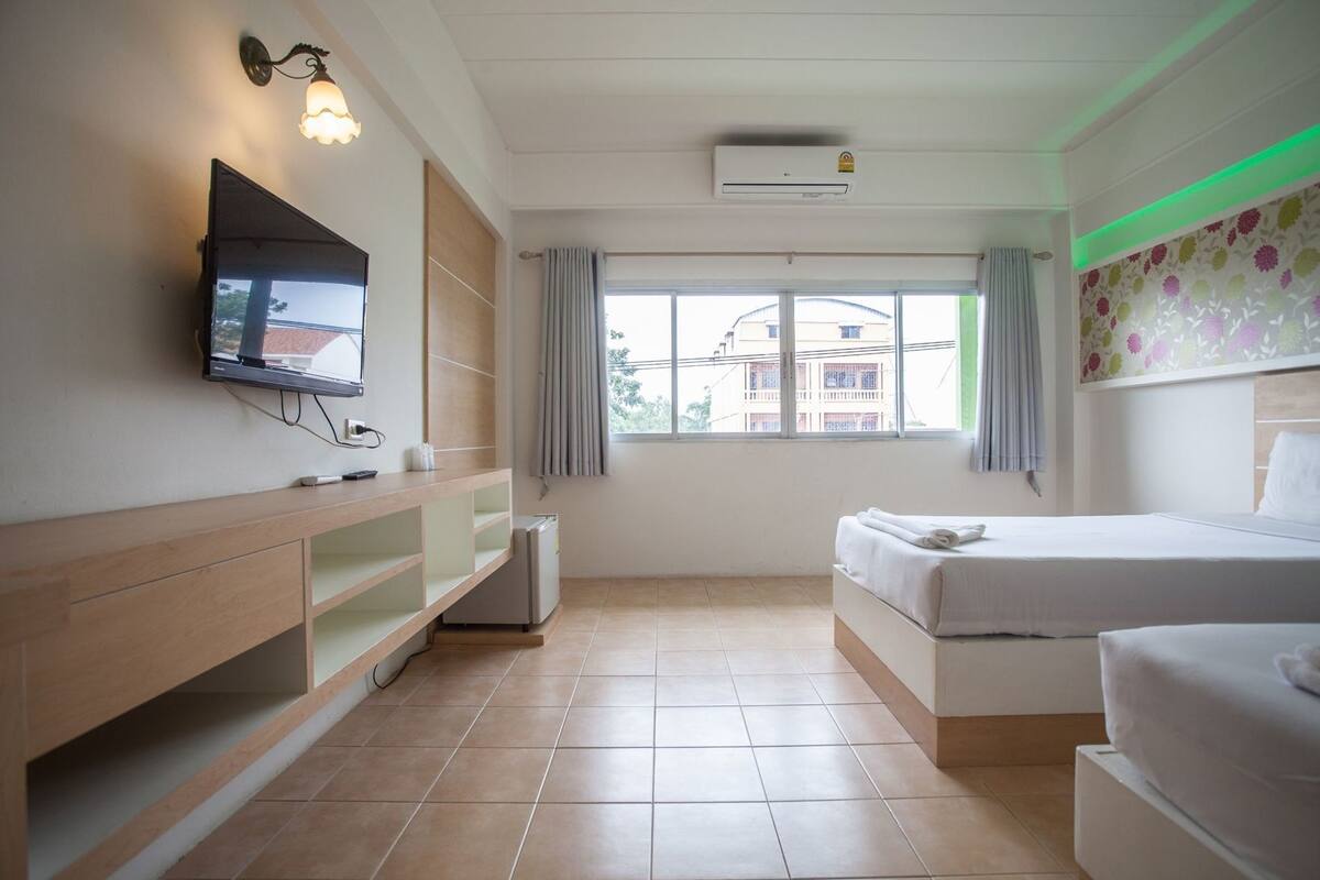 A hotel room with a bed, a TV mounted on the wall, a desk with a chair, a window with curtains, and an air conditioning unit.