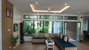 A modern living room with a wooden floor, a green sofa with colorful cushions, a white coffee table, and large windows with a view of greenery. - The Greenery Hotel (Krabi)