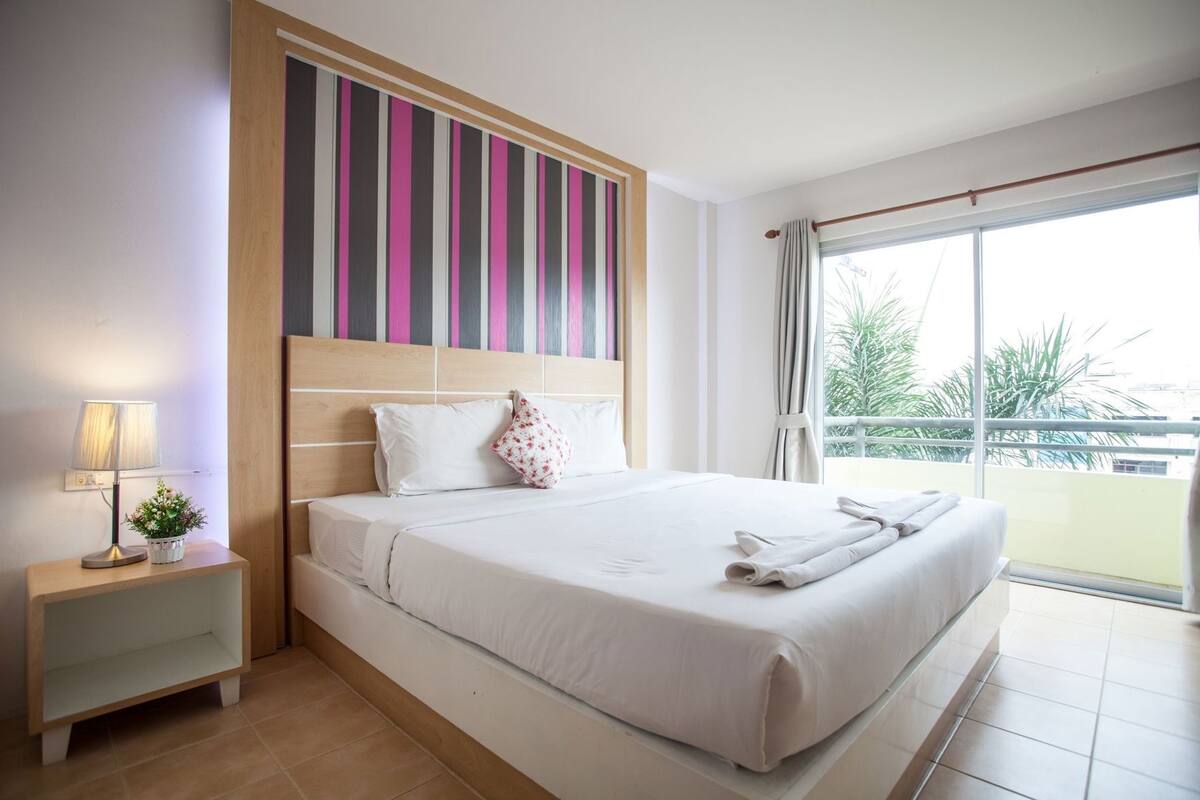 A hotel room with a bed, a nightstand, a lamp, a potted plant, and a view of greenery through the window.