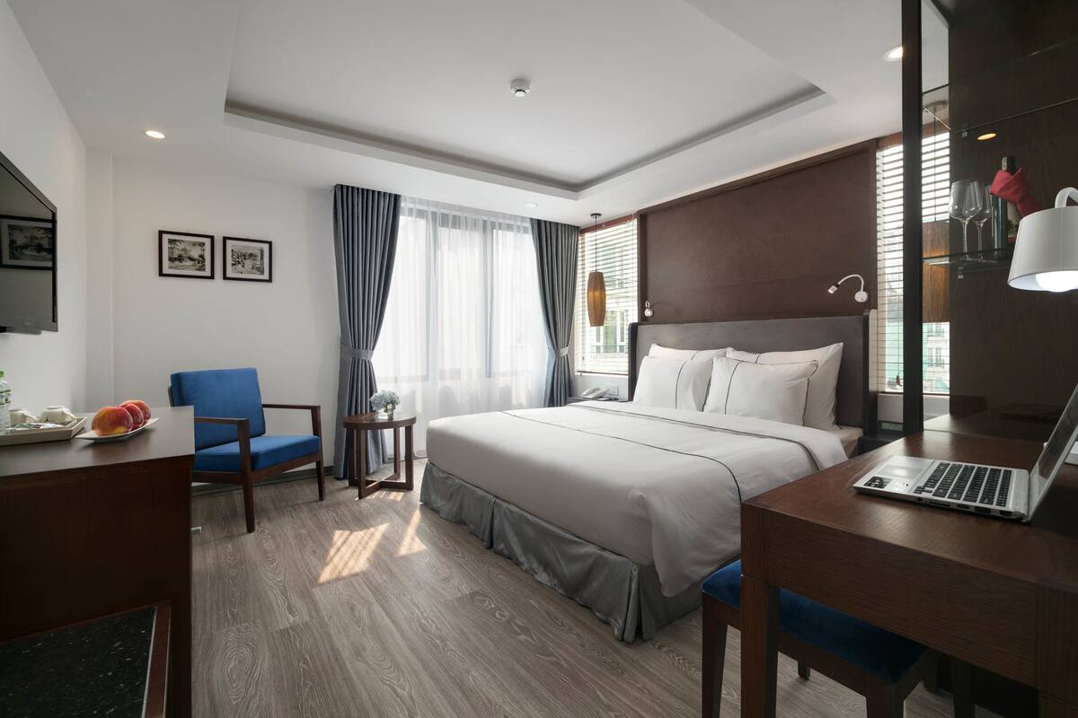 A modern hotel room with a large bed, a desk with a laptop, a chair, and a small table with fruit.