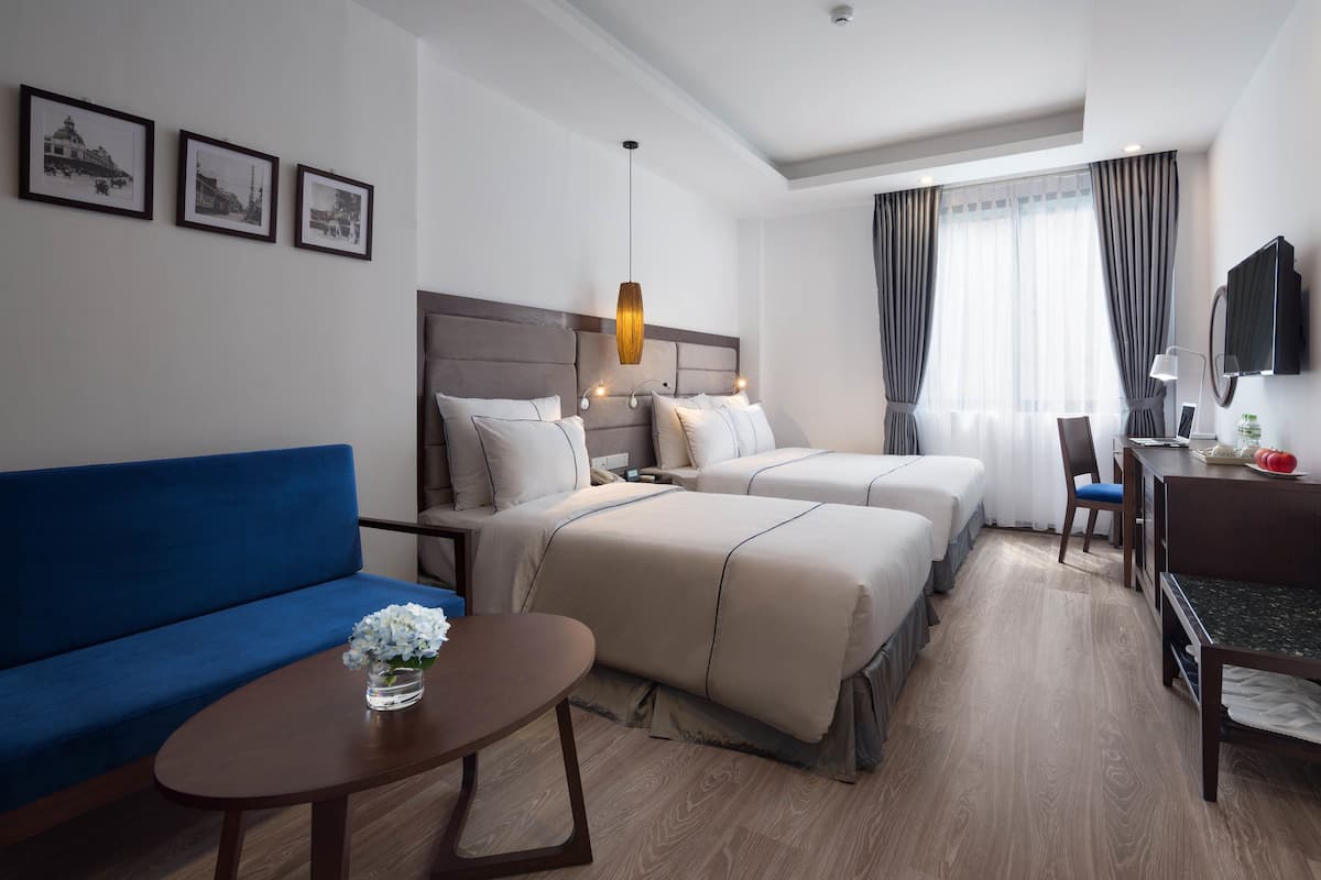 A hotel room with a blue sofa, two beds with beige bedding, a small table with a vase of flowers, a desk with a chair, and a TV mounted on the wall.