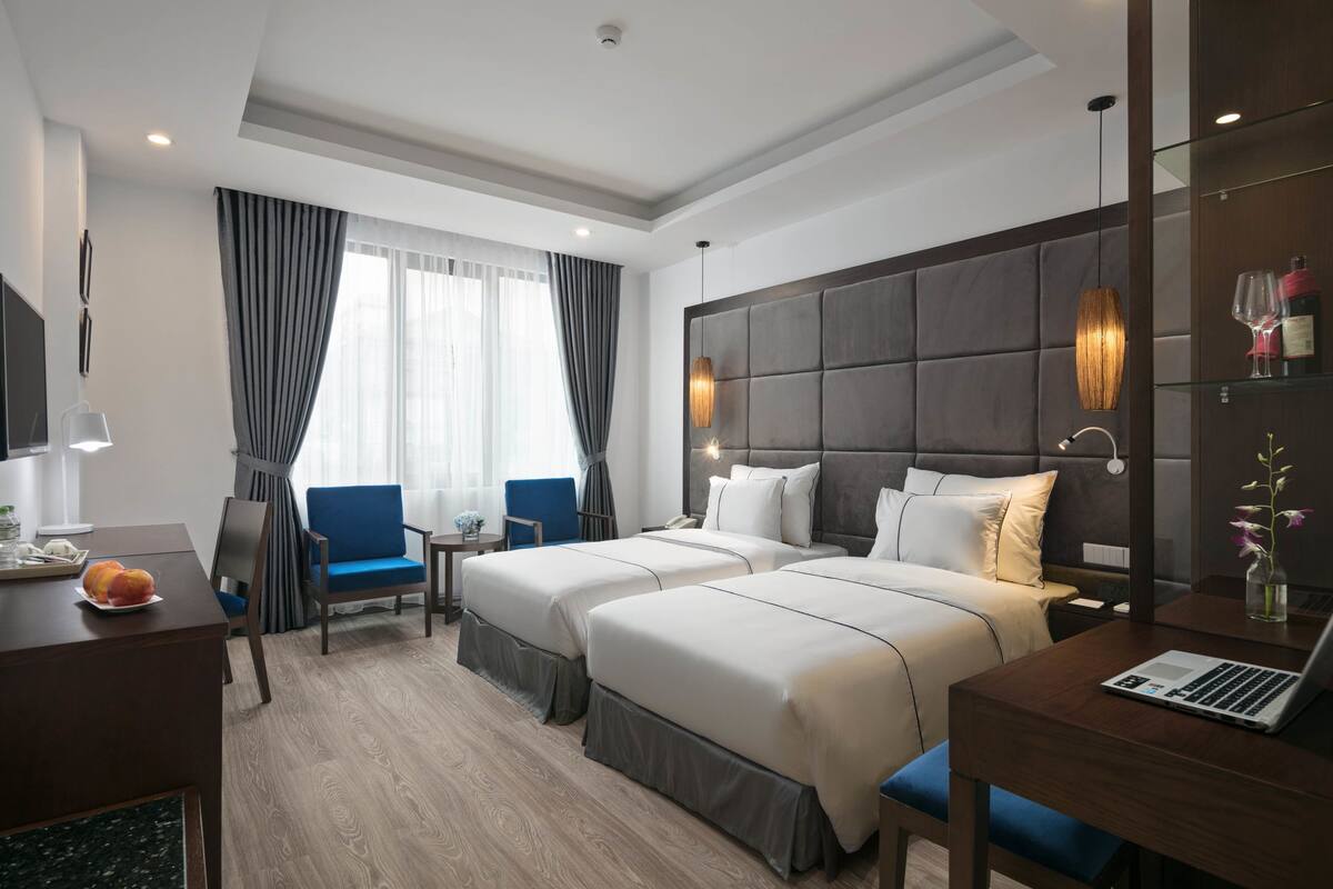A hotel room with two beds, a desk with a laptop, a chair, a TV, and a window with curtains.