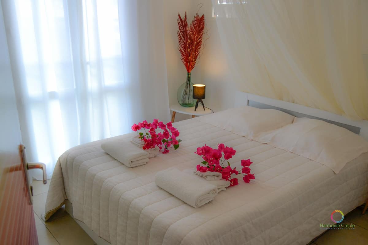 Un lit bien fait, avec du linge de lit blanc et des fleurs roses, une table de chevet avec une lampe et un vase rempli de fleurs rouges.