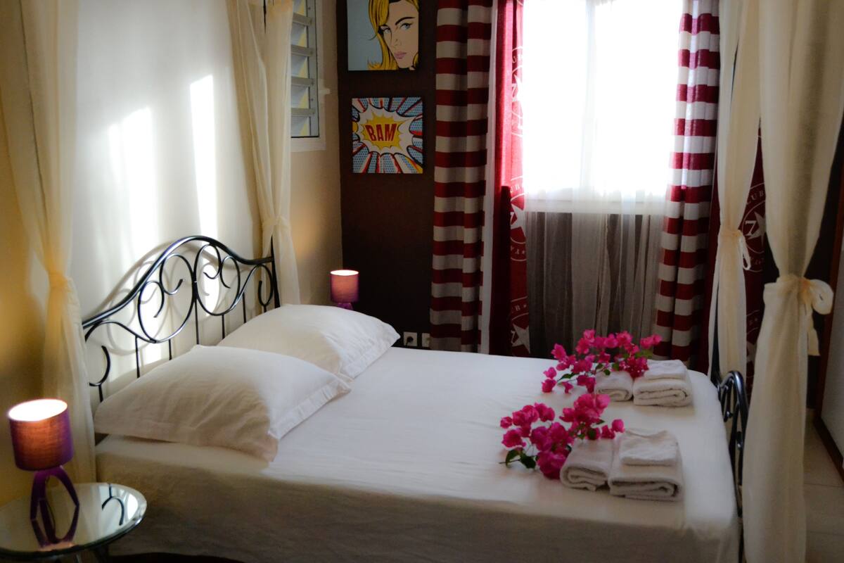 Un lit bien fait, avec du linge de lit blanc, des fleurs roses et des serviettes pliées. On y trouve une table de chevet avec une lampe et un tableau accroché au mur.