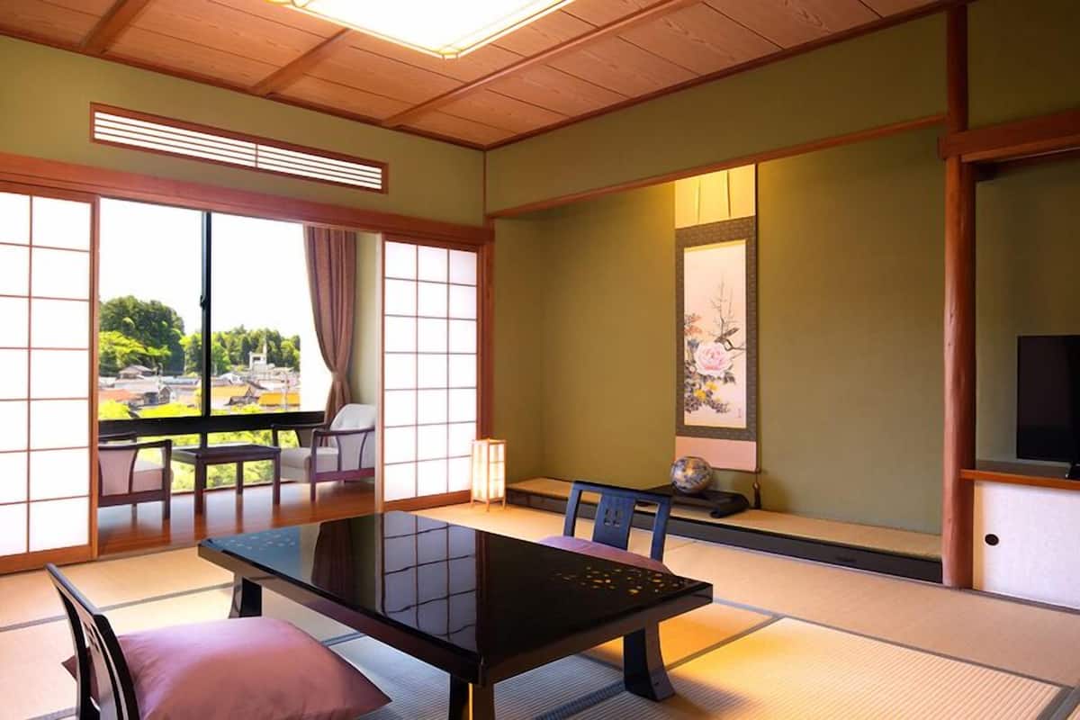 Ein traditioneller japanischer Raum mit Tatami-Boden, einem niedrigen Tisch, einem Fernseher und einer Schiebetür zu einem Außenbereich.