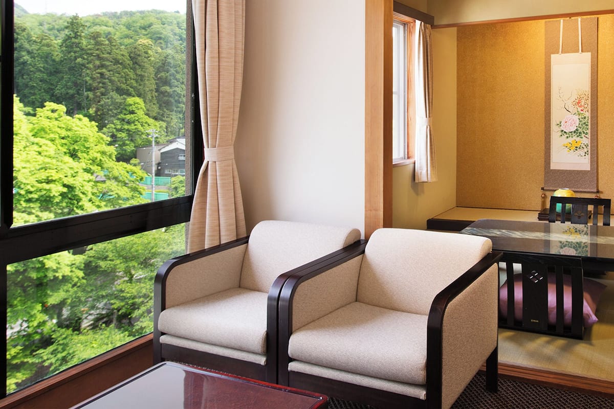 A room with a view of lush greenery, a traditional tatami mat floor, a low table with a cushion, and a chair with a dark frame.