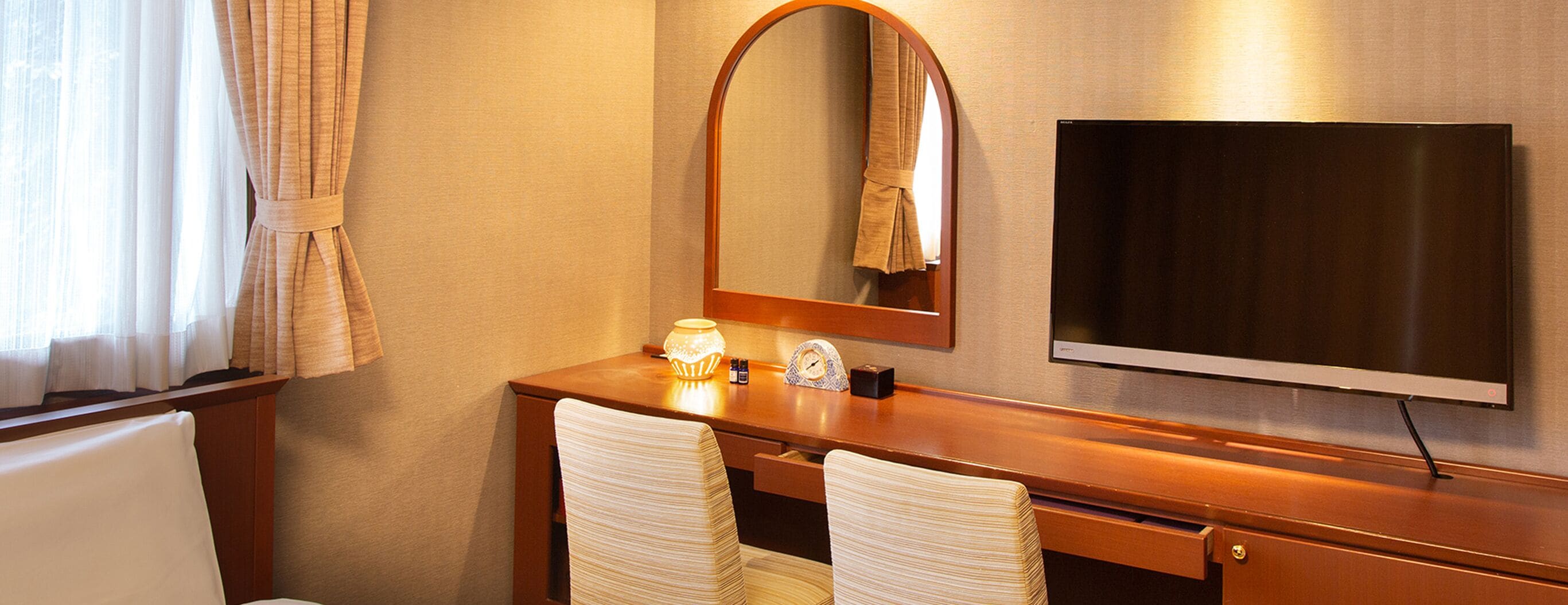 Une chambre d’hôtel avec un bureau en bois, deux chaises, un grand miroir, un téléviseur à écran plat et une fenêtre avec des rideaux.