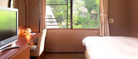 A hotel room with a bed, a desk with a television, and a window with a view of trees and buildings.