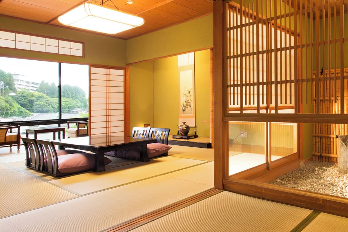 A traditional Japanese room with tatami flooring, a low table with cushions, and a view of greenery outside.