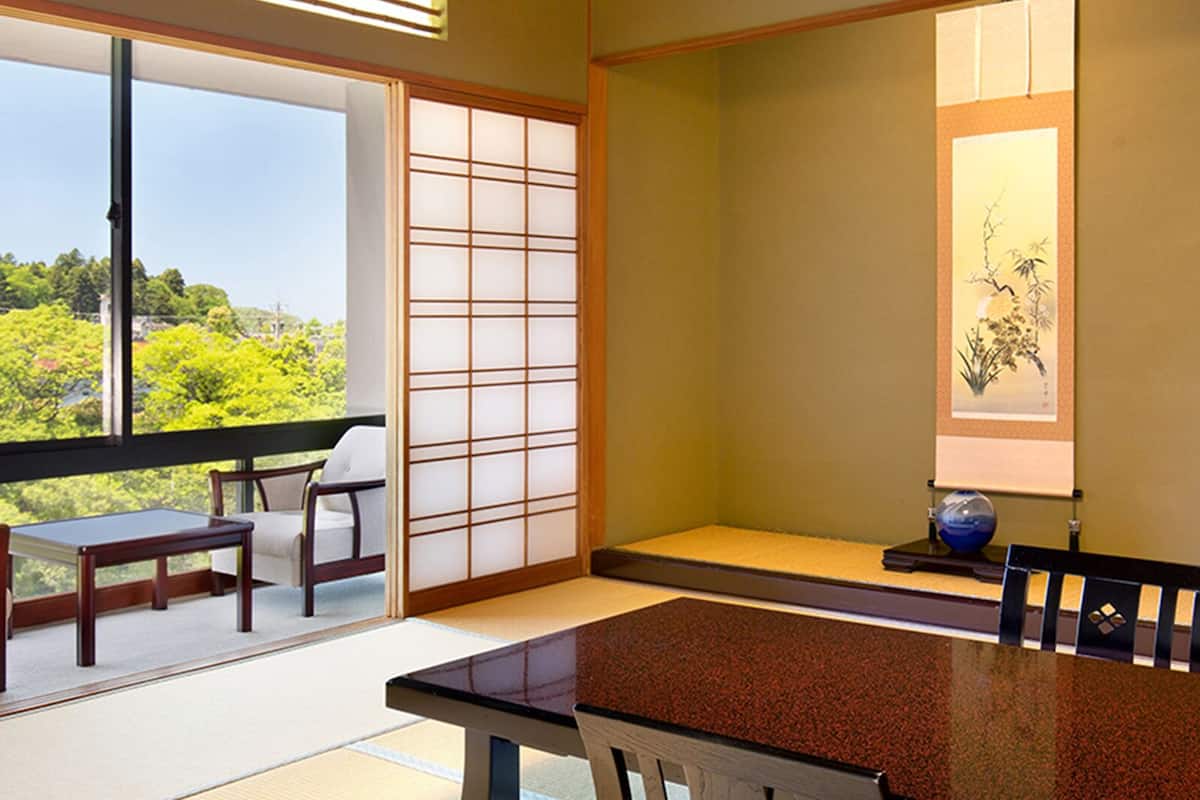 A traditional Japanese room with tatami flooring, a low table, and a sliding door leading to a balcony with a view of greenery.