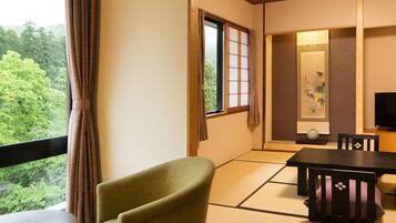 Una habitación japonesa tradicional con suelo de tatami, una mesa baja con cojín, una silla verde, un televisor y vistas a exuberante vegetación.