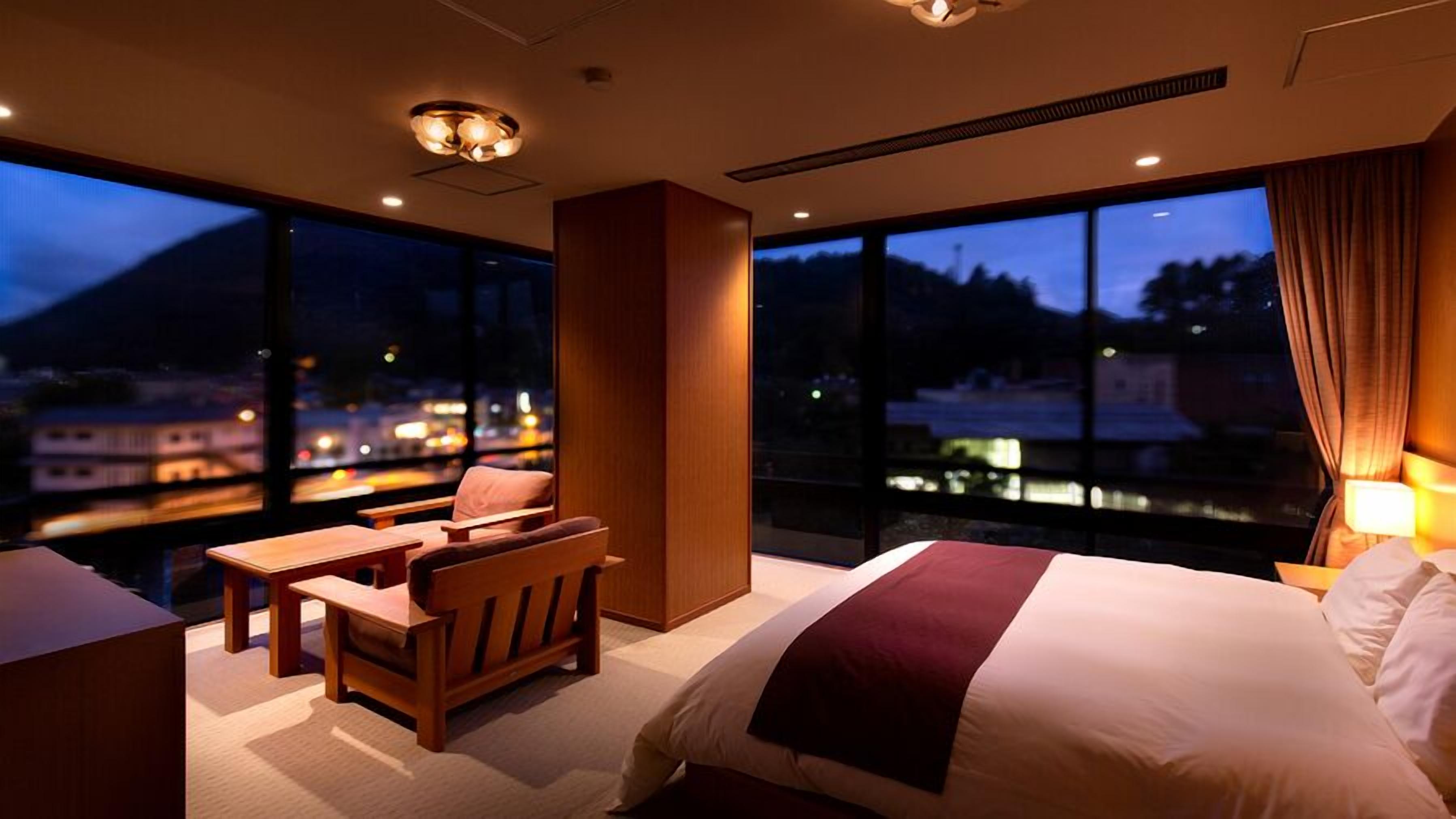 Ein Hotelzimmer mit Bett, Schreibtisch, Stuhl und großen Fenstern mit Blick auf die beleuchtete Stadt bei Nacht.