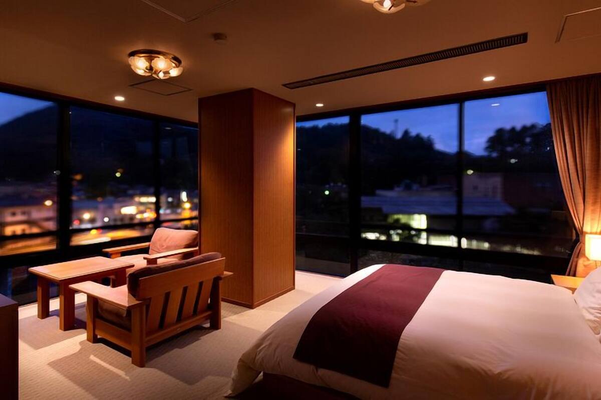 Habitación de hotel con cama, escritorio, silla y grandes ventanales con vistas a la ciudad de noche.