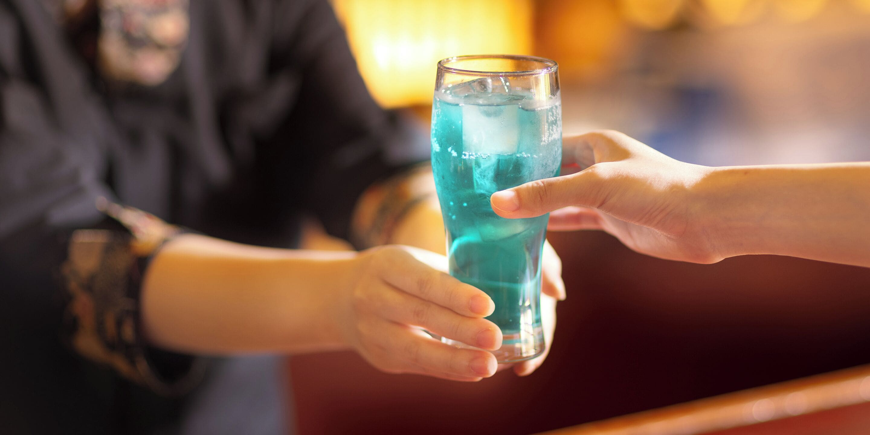 Una persona sosteniendo un vaso alto con líquido azul, probablemente una bebida, en un bar.