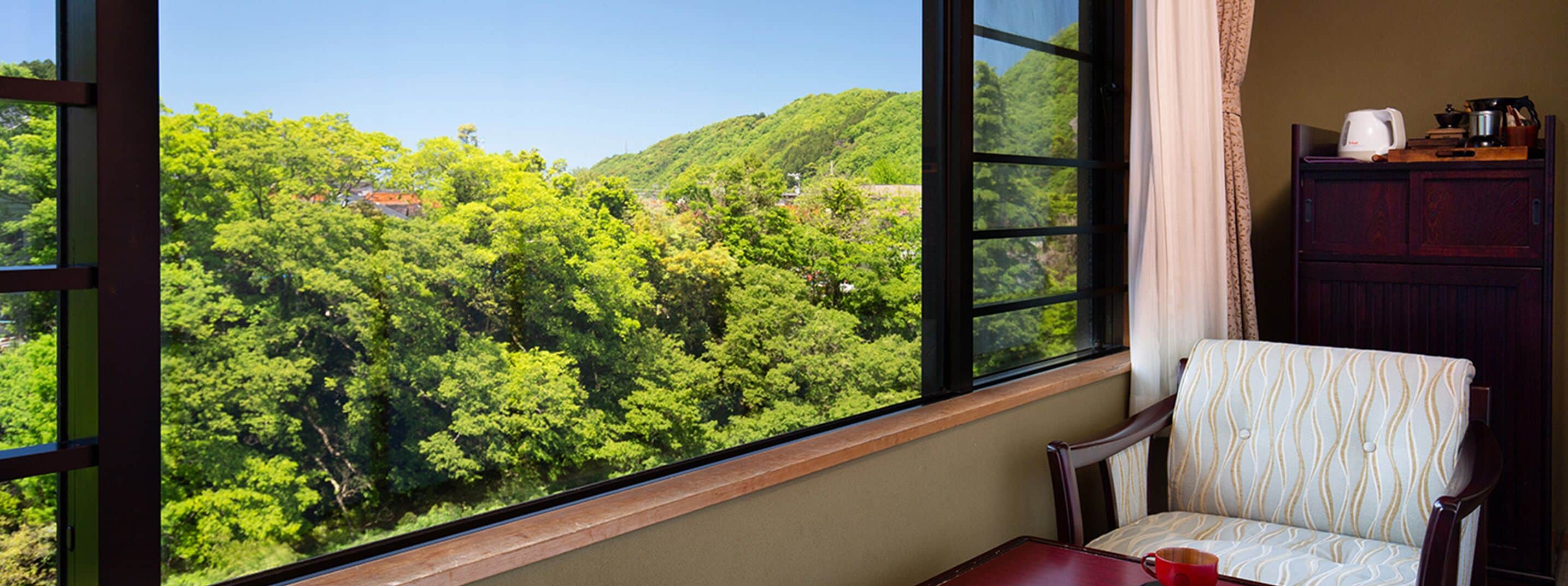 Una habitación con un ventanal grande que ofrece vistas a un bosque verde y frondoso. Hay una silla de madera con un cojín estampado y una mesita con una taza roja.