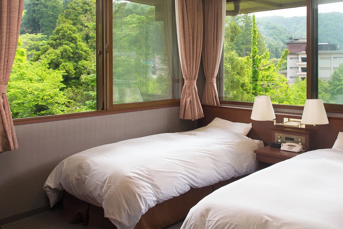 A hotel room with two beds, a nightstand with a lamp, and a view of a forested mountain area.