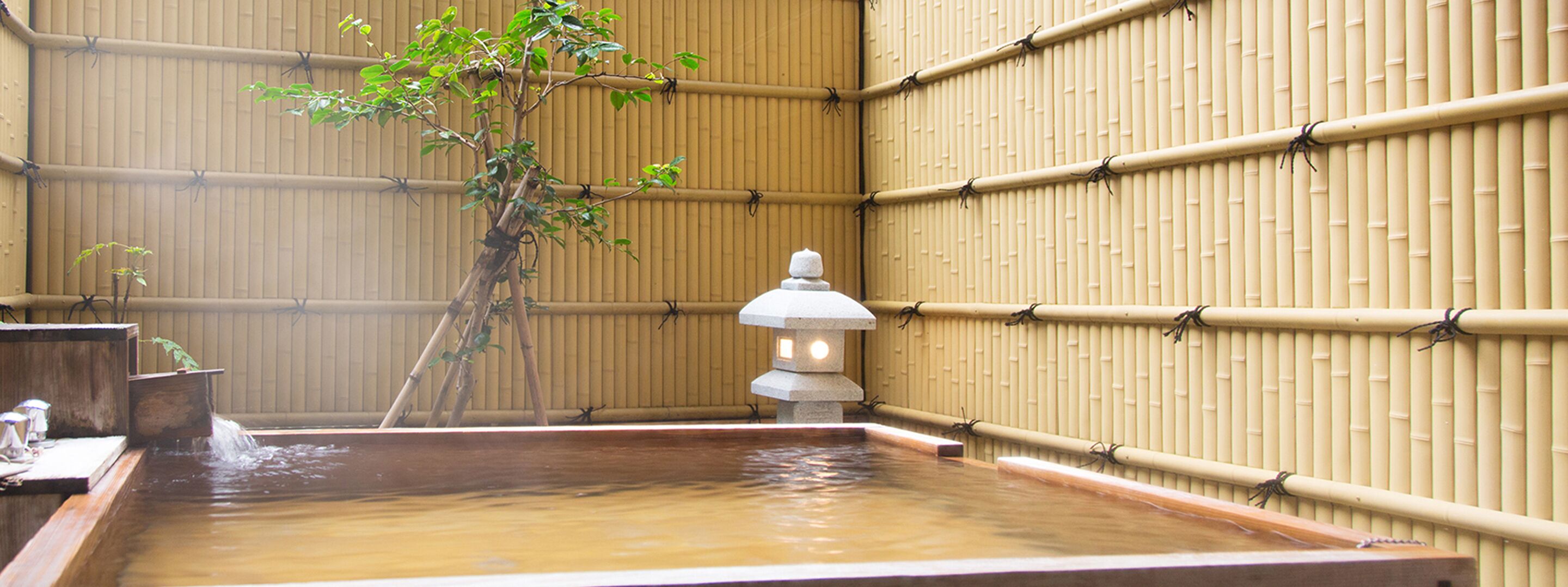 Uma onsen japonesa tradicional com uma fonte de pedra, paredes de bambu e uma pequena árvore.