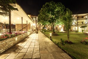 A well-lit pathway leading to a building with multiple balconies and a garden area with flowers and a lawn. - The Lakeside Retreat Pokhara by IME Hospitality (Pokhara)