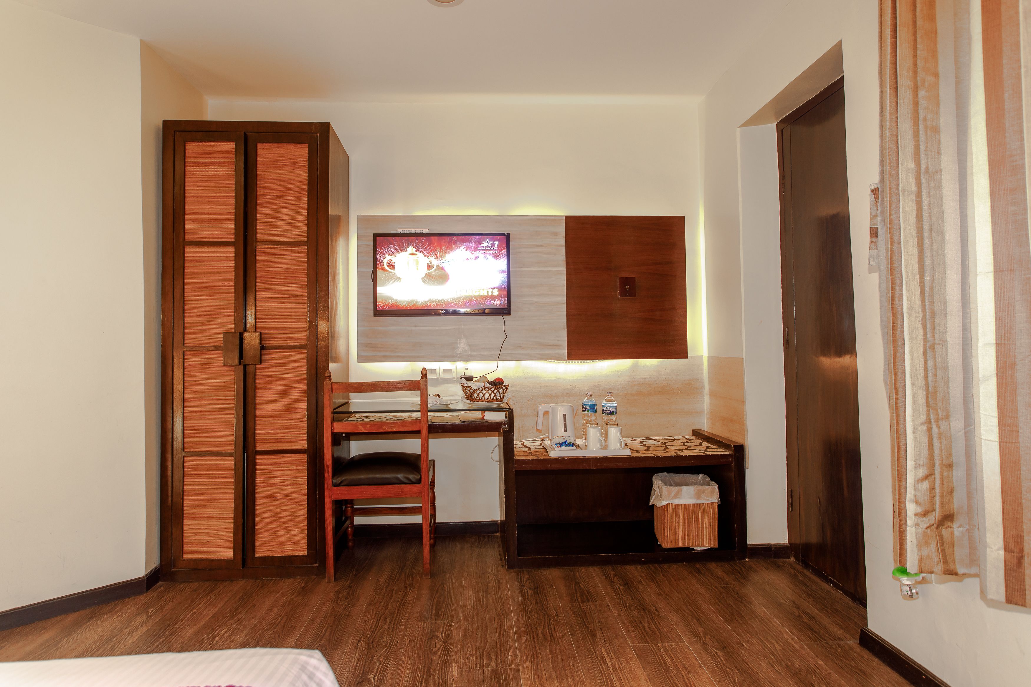 A hotel room with a wooden desk, a television mounted on the wall, a wardrobe, and a small trash bin.