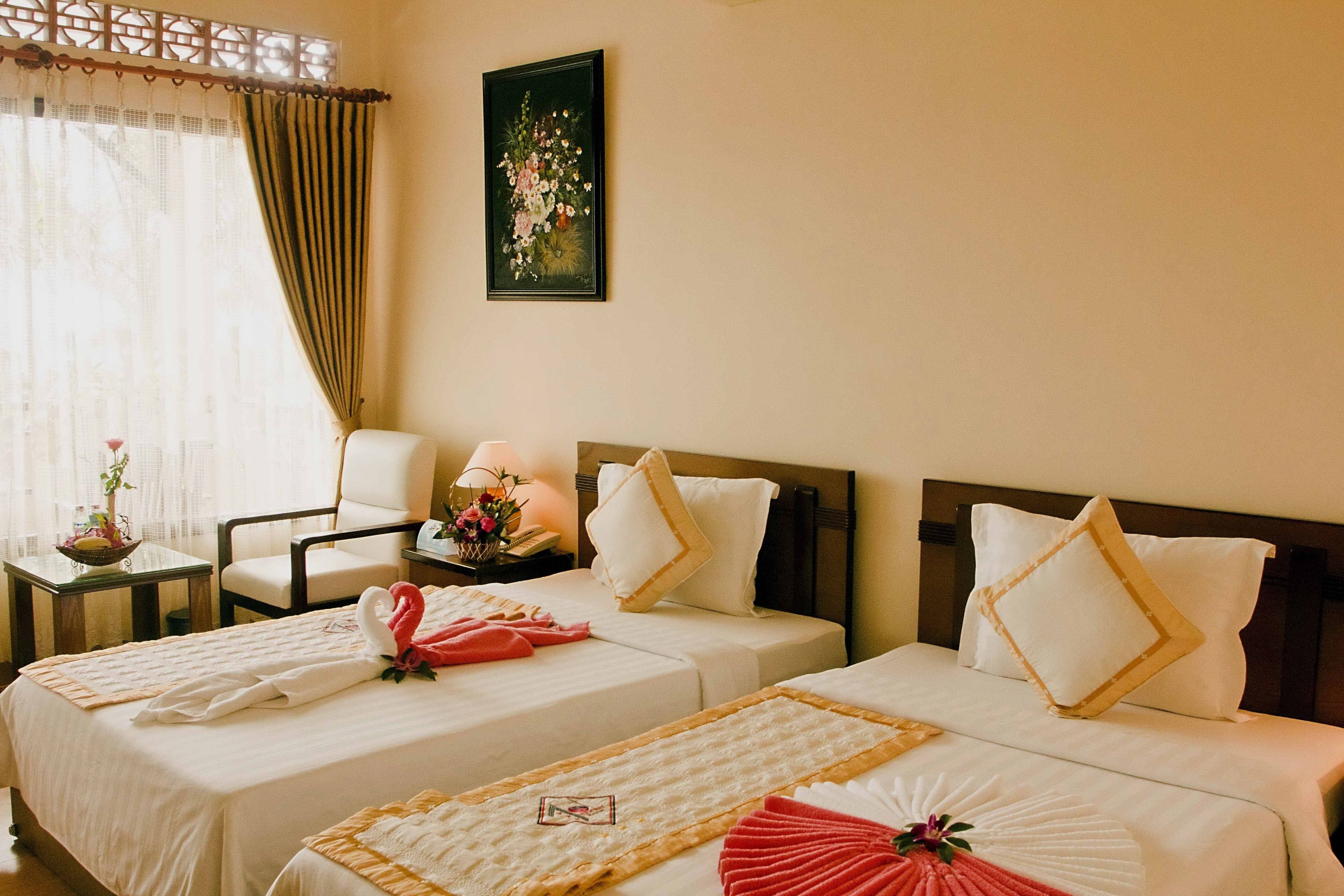 Una habitación de hotel con dos camas, una silla, una mesa con un florero y un cuadro en la pared.
