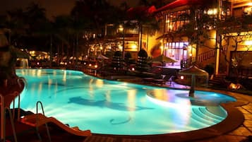 Vista nocturna de un área de piscina de hotel, con una piscina bien iluminada, sillas de descanso y un edificio con varios balcones.