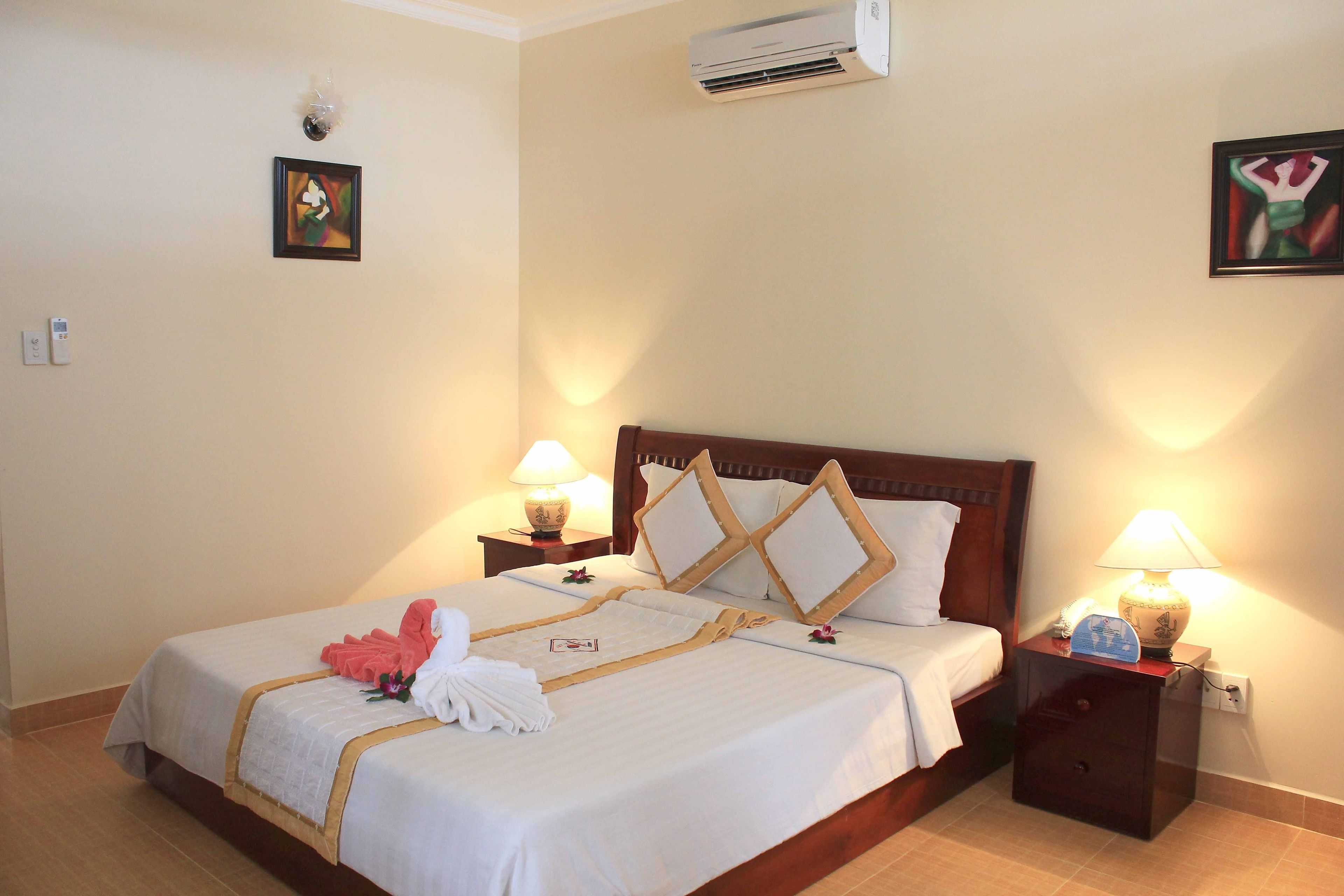 A neatly made bed with white linens and decorative pillows, a bedside table with a lamp, and a wall-mounted air conditioner.