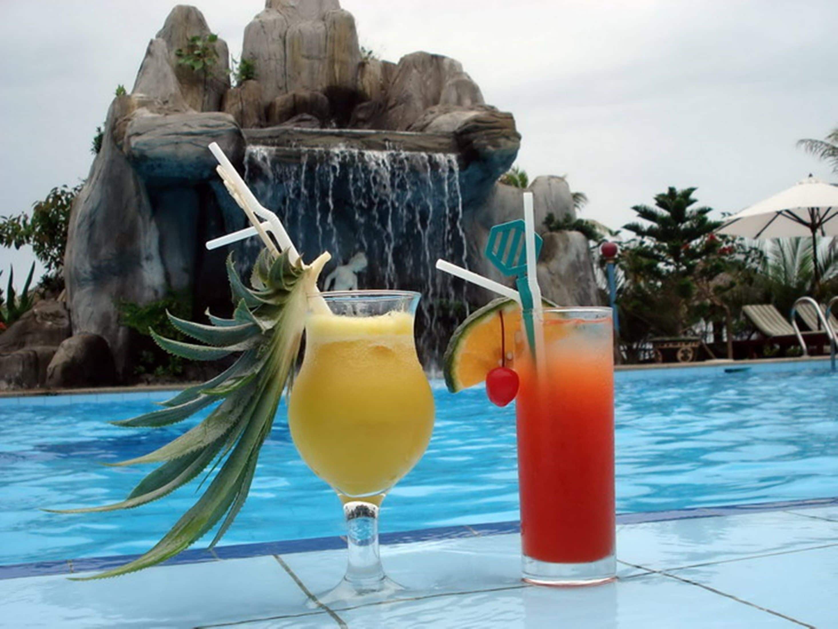 Zona de piscina con una cascada y dos bebidas, una adornada con piña y la otra con cereza.