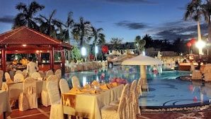 Un área de comedor junto a la piscina con mesas dispuestas para un evento formal, rodeada de palmeras y un cielo despejado.