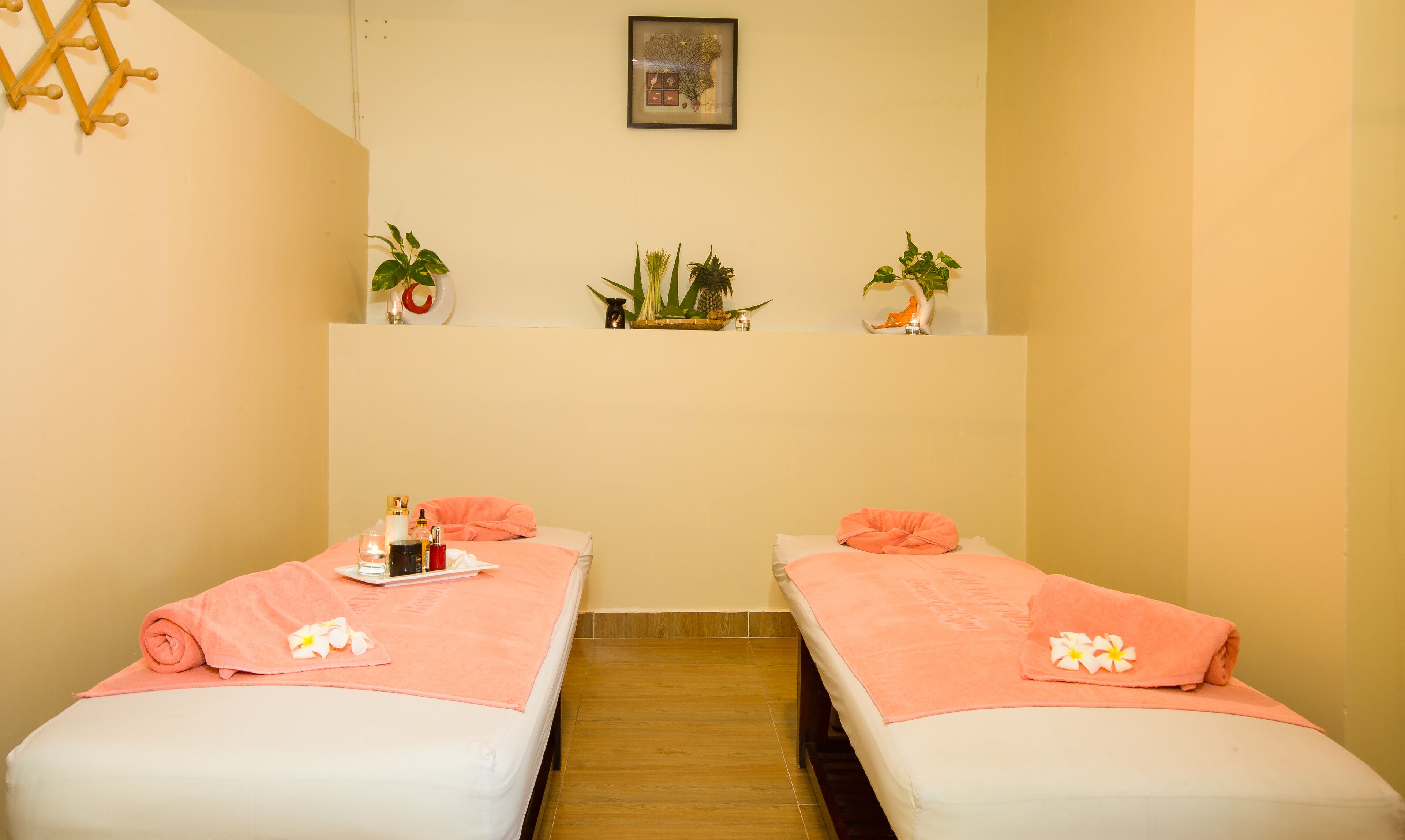 Une salle de spa avec deux lits, chacun agrémenté d’une serviette rose et d’une fleur. Des étagères au-dessus des lits accueillent des plantes et un tableau encadré.