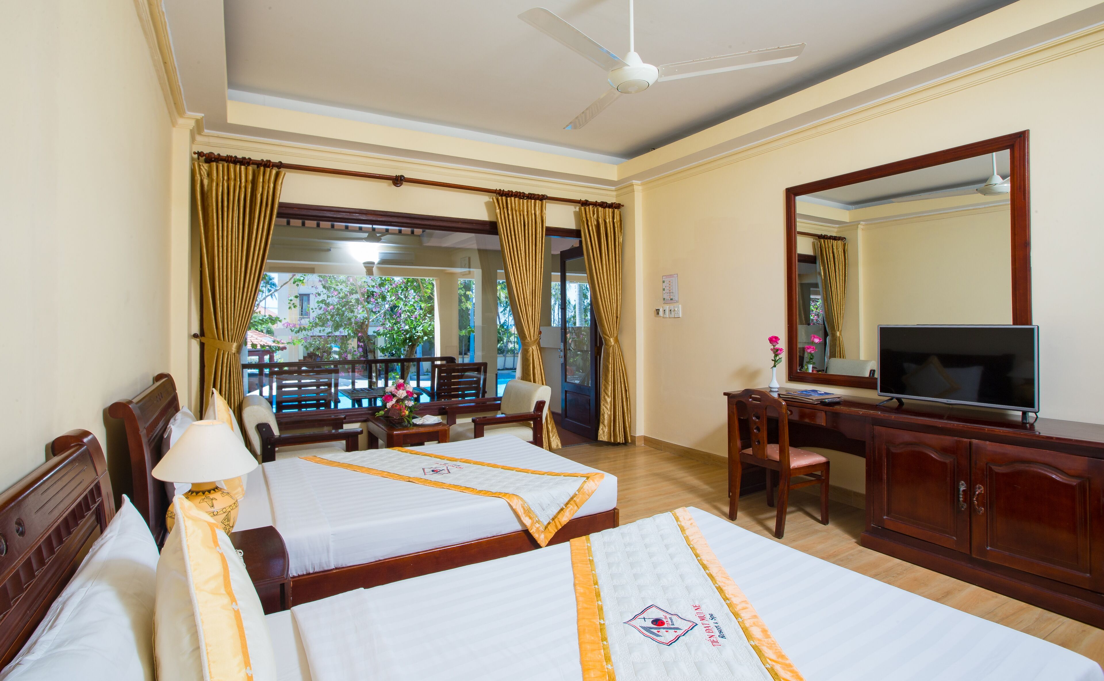 A hotel room with two beds, a desk with a TV, a chair, a desk lamp, and a view of the outside.