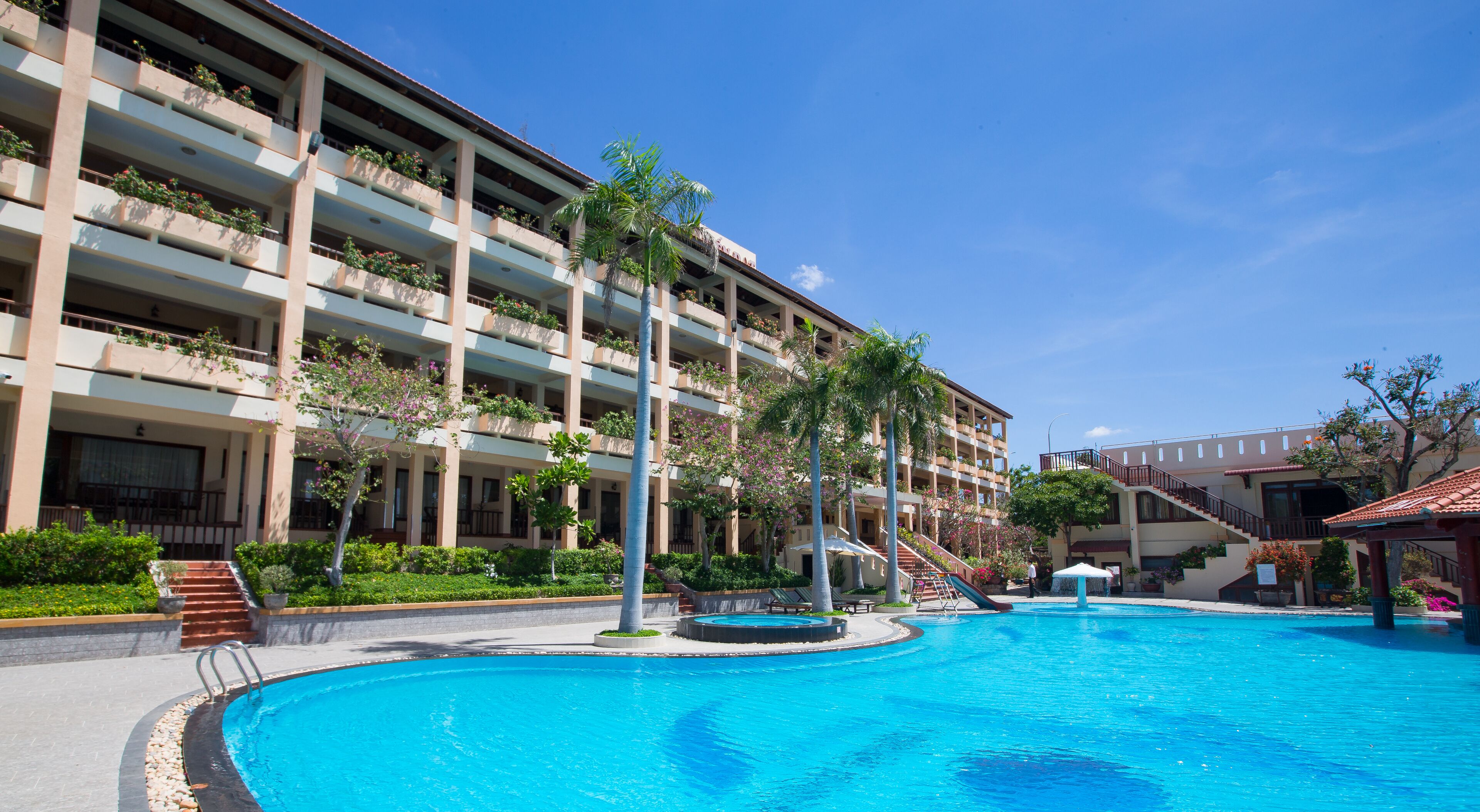 Un hotel con una gran piscina, varios balcones con plantas y un cielo azul despejado.
