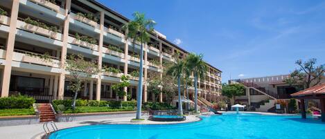Un hotel con una gran piscina, varios balcones con plantas y un cielo azul despejado.