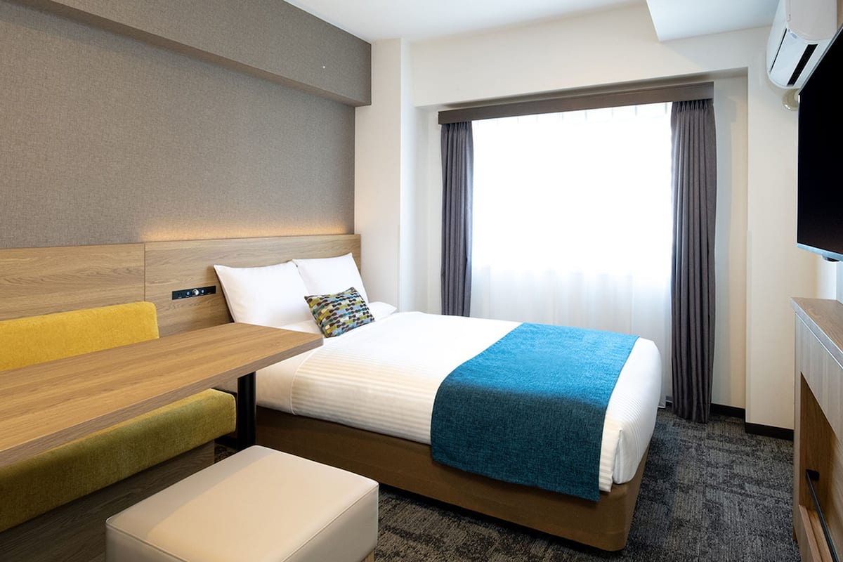Quarto de hotel moderno com uma cama bem arrumada, uma escrivaninha com cadeira, um pequeno sofá e uma televisão.