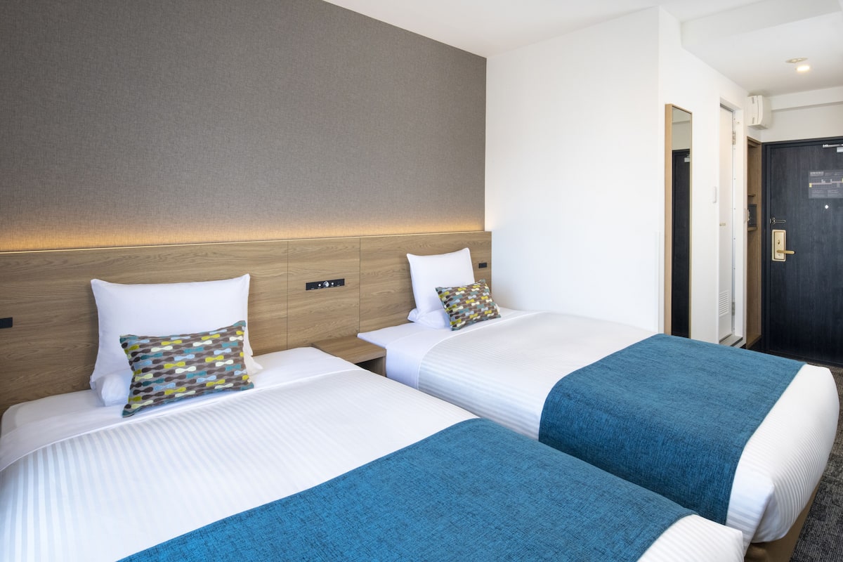 Quarto de hotel com duas camas, cada uma com colchão branco e manta azul. Possui cabeceira de madeira, mesa de cabeceira pequena e luminária fixada na parede.