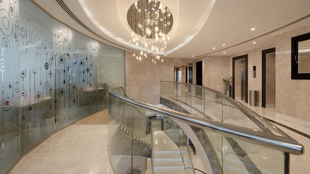 A modern interior with a glass railing staircase, a decorative wall with circular patterns, and a central chandelier. - Landmark Premier Hotel (Dubai)