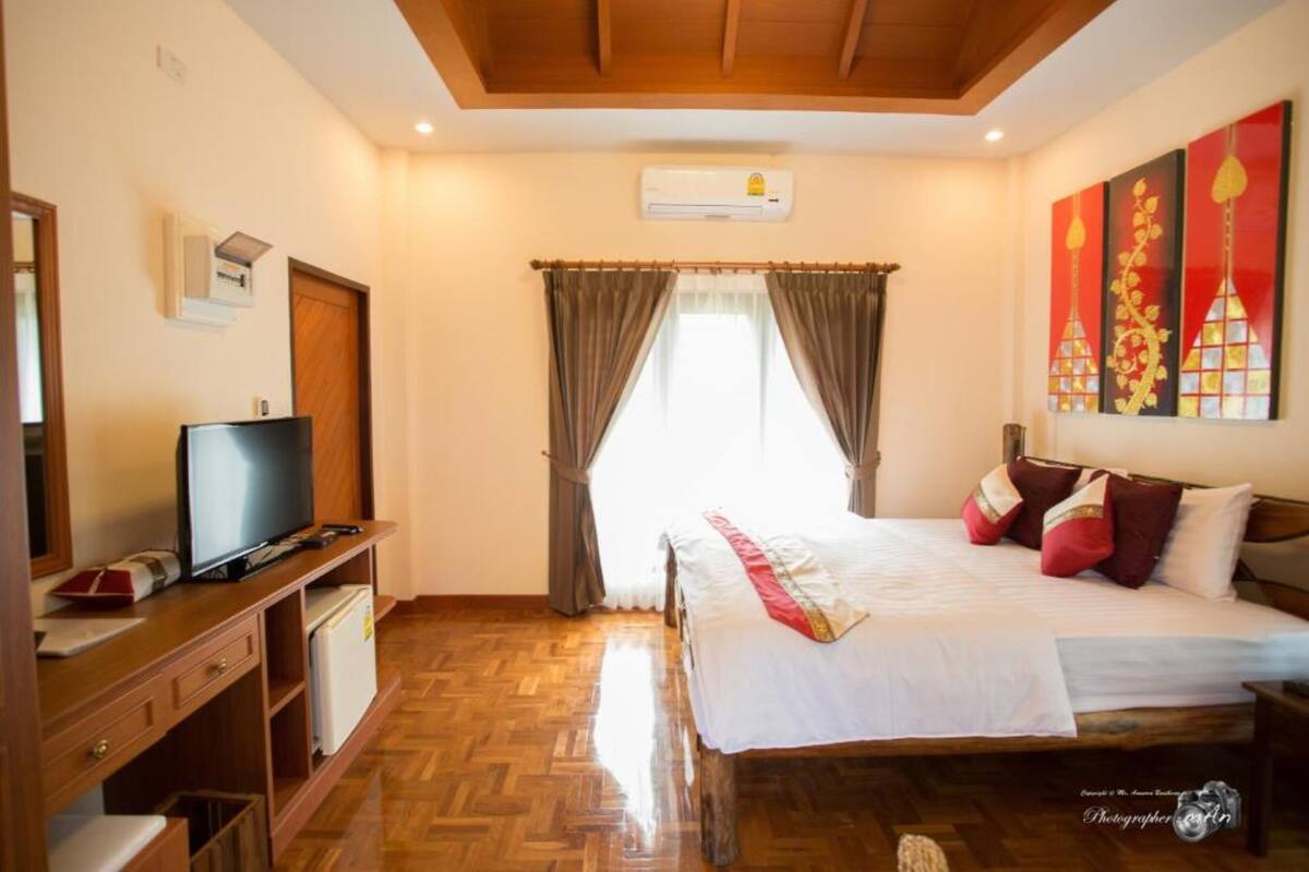 A bedroom with a wooden bed, a TV on a wooden cabinet, a wardrobe, and a window with curtains.