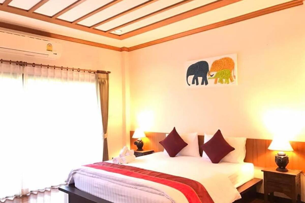 A hotel room with a bed, two bedside tables, a nightstand, a window with curtains, and a wall with a picture of elephants.