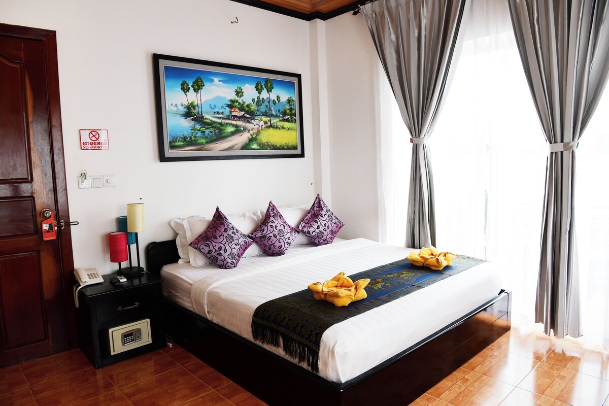 A hotel room with a bed, a nightstand with a phone and lamp, a painting on the wall, and a view of the outdoors through the window.