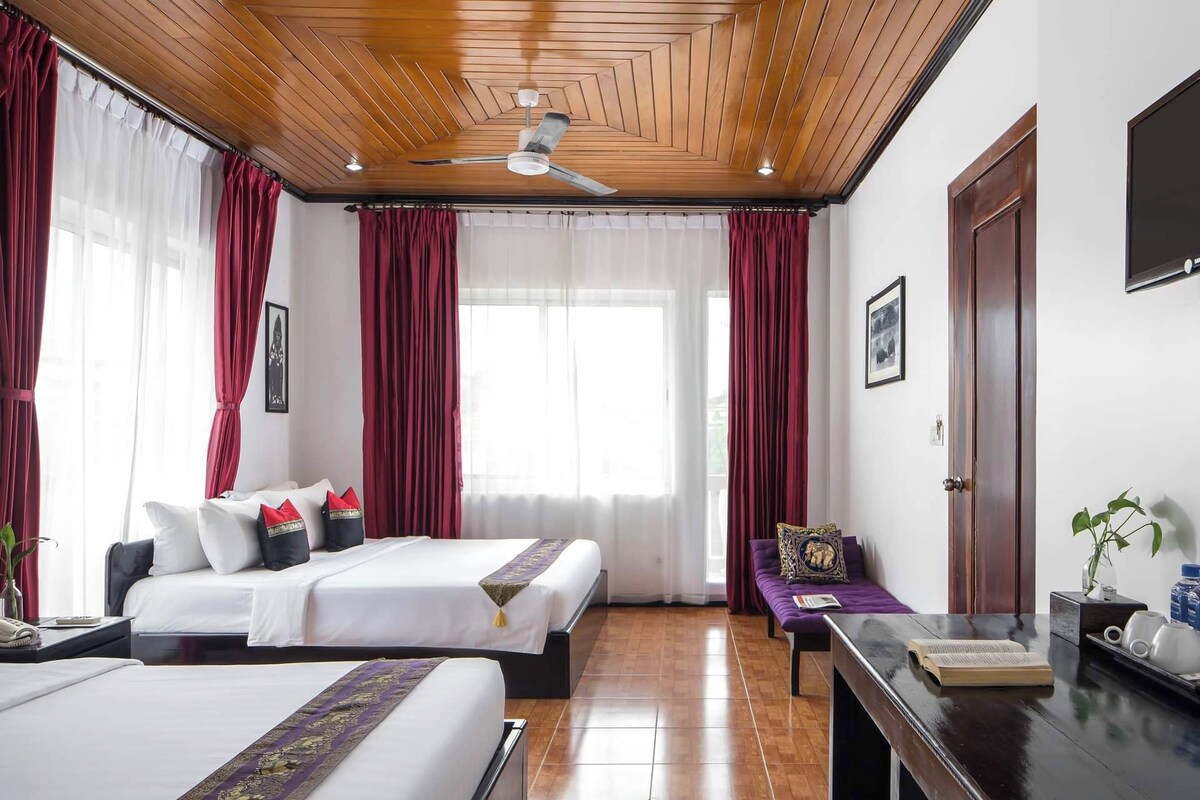 A hotel room with two beds, a ceiling fan, large windows with red curtains, a wooden ceiling, and a TV on the wall.