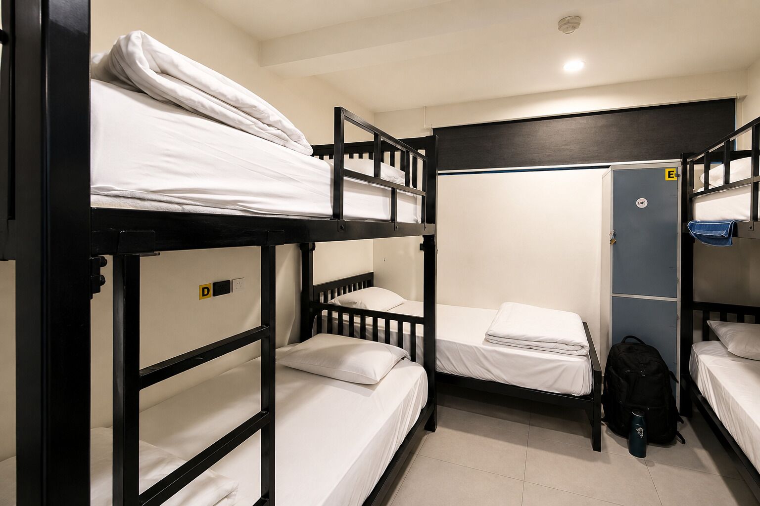 A dormitory room with bunk beds, a wardrobe, and a door marked with an 'E' for emergency exit.