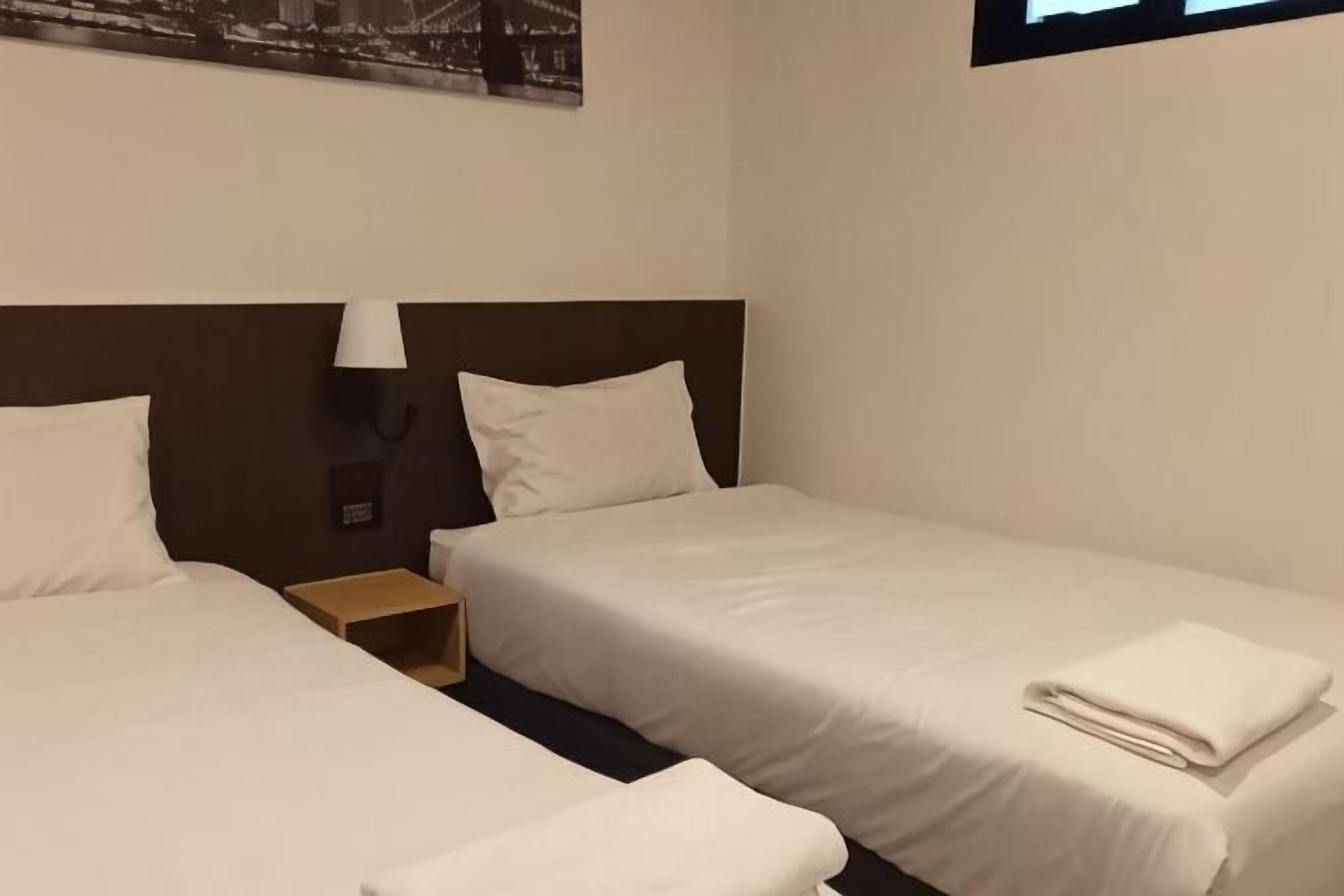 A hotel room with two single beds, a nightstand with a lamp, and a wall-mounted phone.