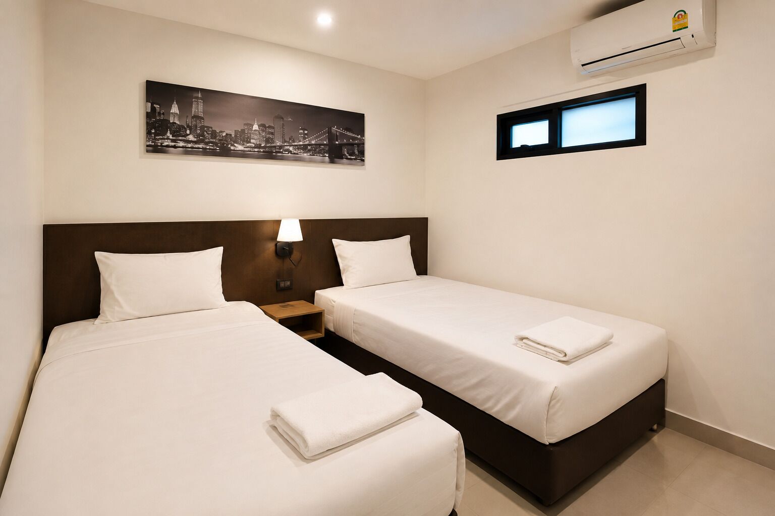 A hotel room with two beds, a nightstand, a lamp, an air conditioning unit, and a cityscape painting.