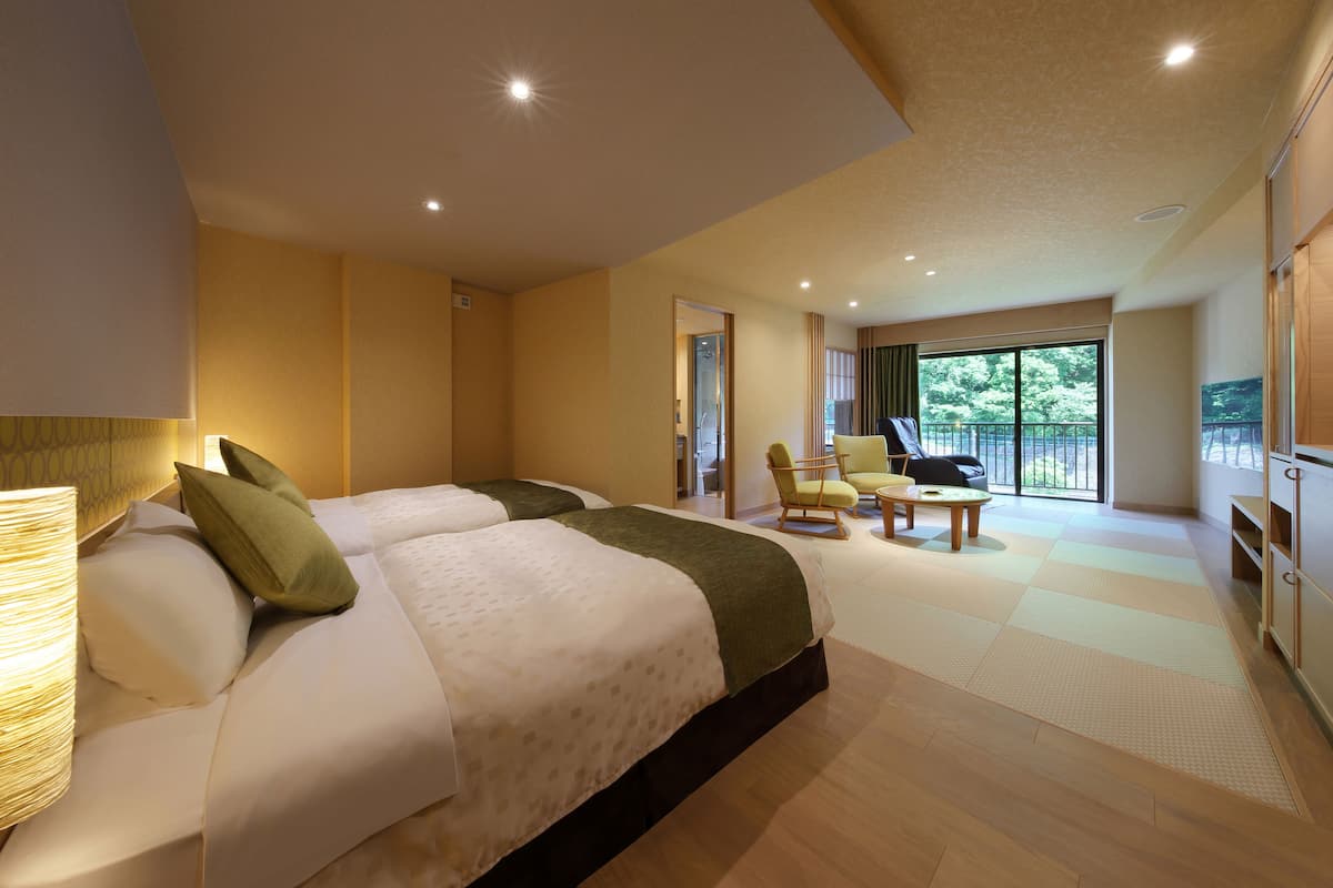 Une chambre d’hôtel avec deux lits, un coin salon avec un fauteuil et une table, et une grande fenêtre donnant sur un paysage verdoyant.