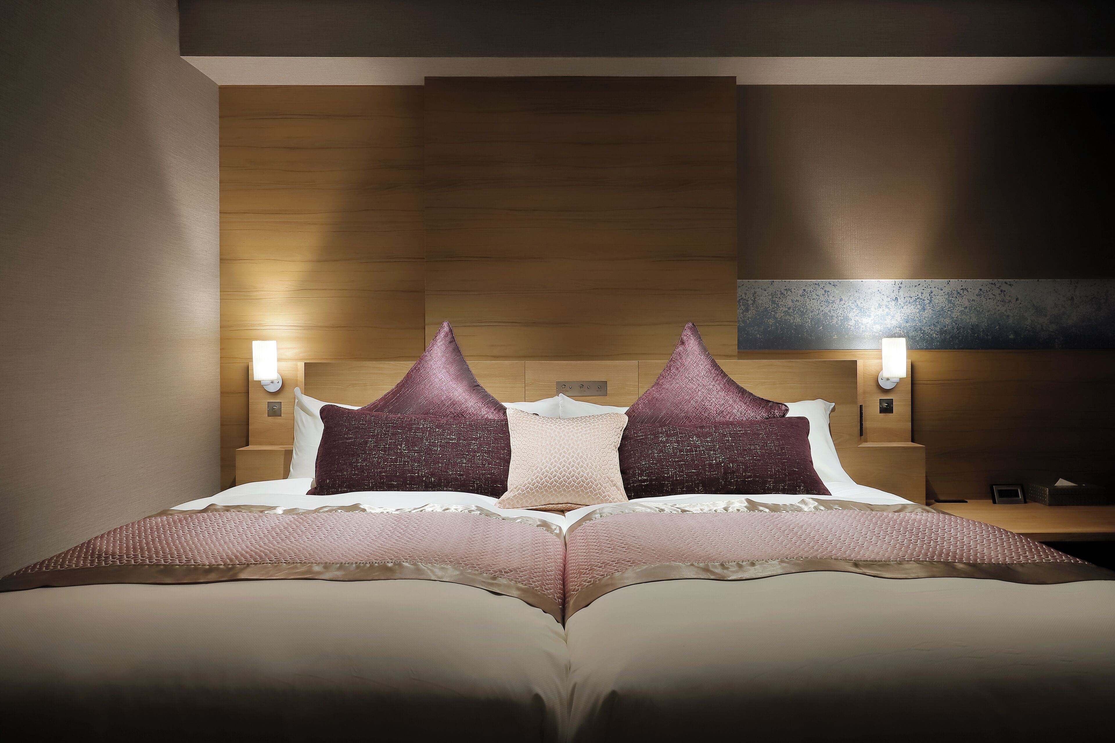 A neatly made bed with a textured blanket, two decorative pillows, and a wooden headboard. There are two wall-mounted nightstands with lamps.