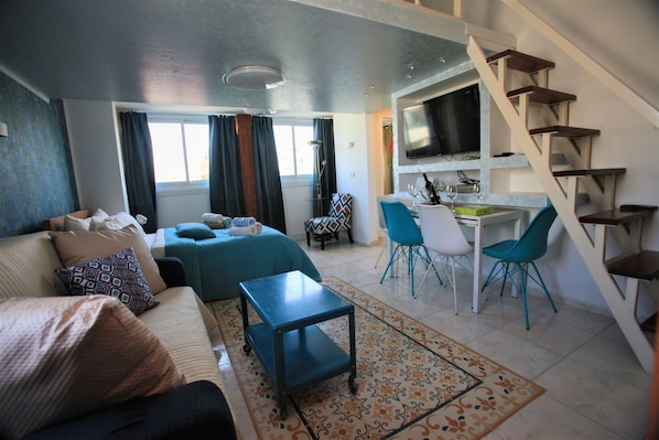 Una sala de estar con sofá, una mesa de centro y un tapete estampado. Se ve una escalera que conduce a un comedor con sillas y un televisor.