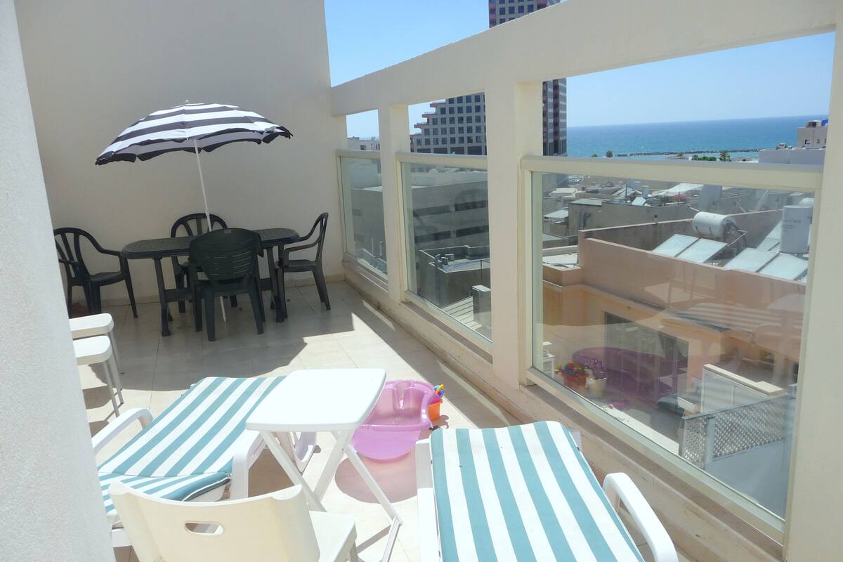 Un balcón con mesa y sillas, un parasol a rayas y vista al mar.