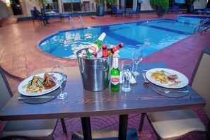 A dining table with two plates of food, a bucket with bottles, and a pool in the background. - Mika Hotel, Kabulonga (Lusaka)